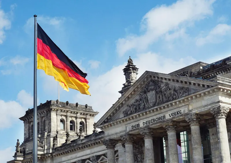 Bandera alemana ondeando frente a la fachada del edificio del Reichstag bajo un cielo azul.