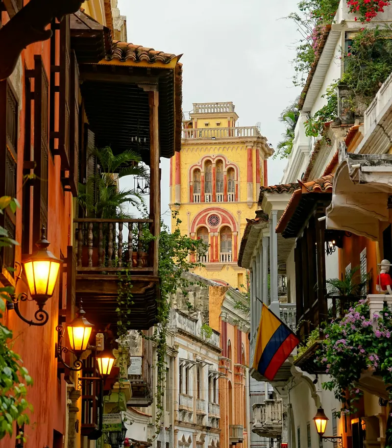 Calle estrecha en Cartagena con balcones coloniales de madera y la torre del reloj amarilla al fondo.