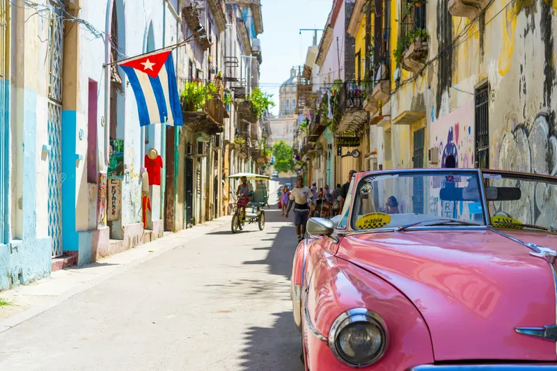 Auto clásico rosado estacionado en una calle adoquinada de La Habana con edificios coloridos y una bandera cubana.
