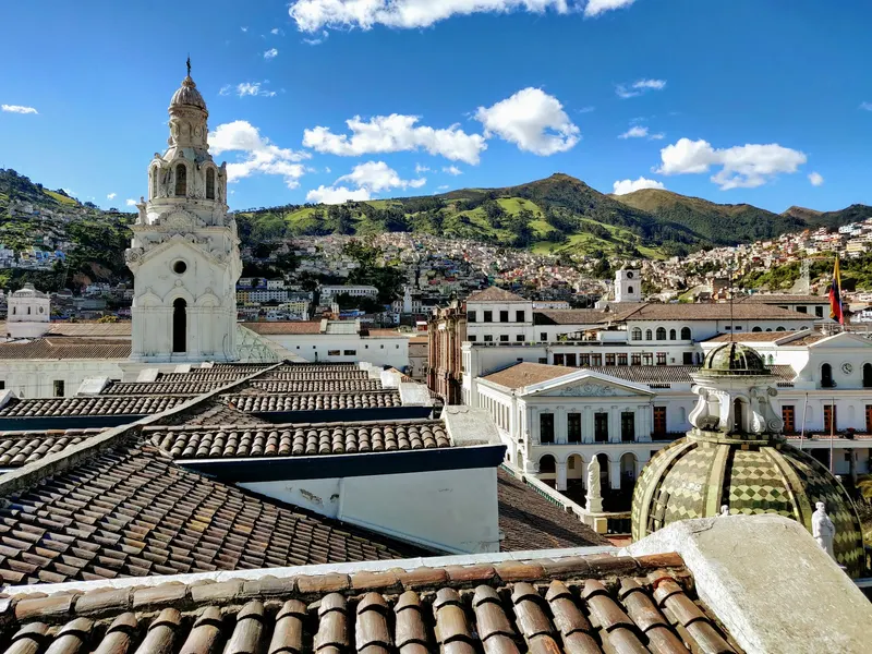 Vista de los tejados del centro histórico de Quito con la torre blanca de una iglesia y el cerro Panecillo de fondo.