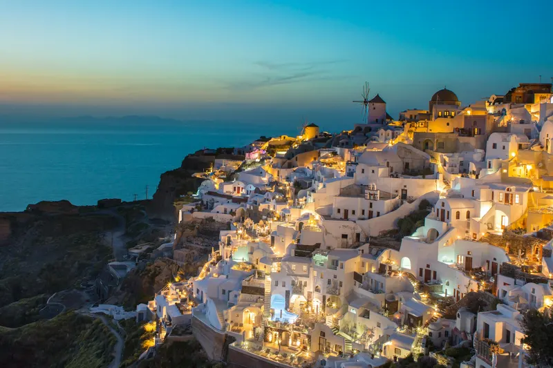 Casas blancas con cúpulas y molinos de viento iluminados al anochecer en un acantilado de Santorini junto al mar.