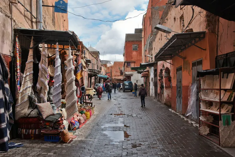 Calle estrecha en Marrakech con coloridos tejidos y lámparas colgando fuera de las tiendas con paredes rojizas.