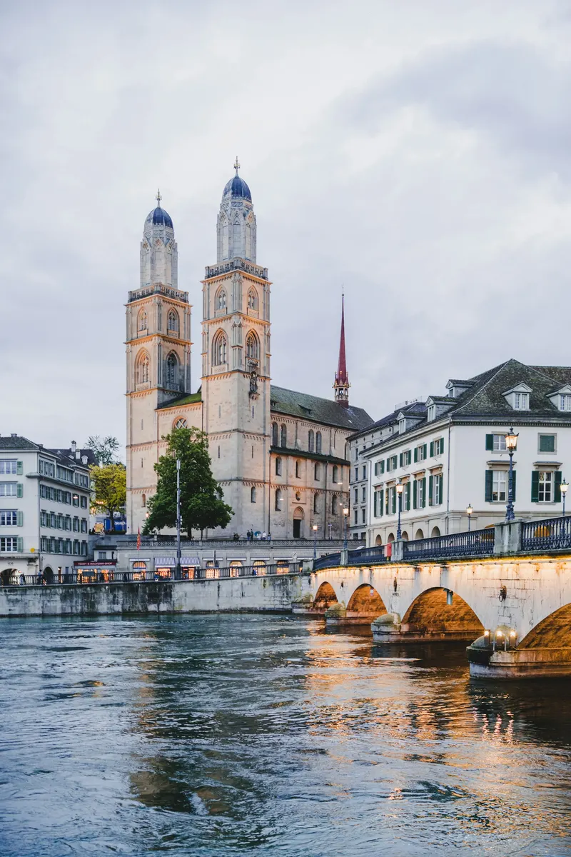 Iglesia de Grossmünster con sus dos torres destacando sobre el río Limmat y un puente de piedra iluminado al atardecer.