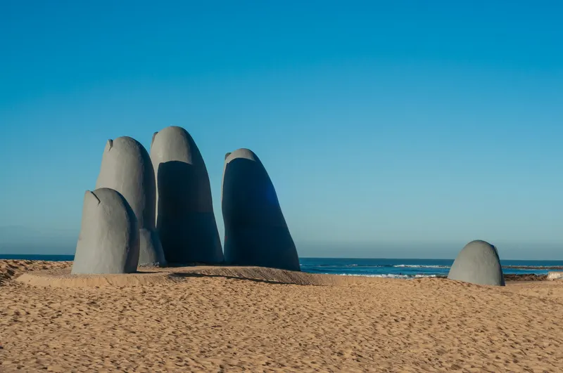 Escultura de piedra con forma de dedos emergiendo de la arena en una playa con el océano y el cielo azul de fondo.